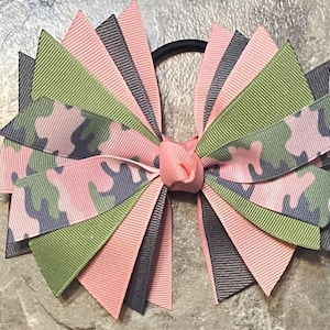 May include: A pink, green, and gray camouflage ribbon hair bow with a black elastic hair tie.