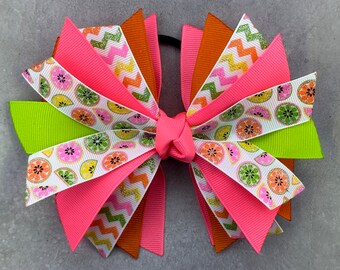 May include: A colourful hair bow with a lime, lemon, and grapefruit print. The bow is made of pink, green, orange, and white ribbon.