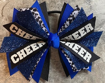 May include: A blue and black cheer bow with white glitter and the word "CHEER" on each side. The bow is tied with a blue ribbon and has a black elastic band.
