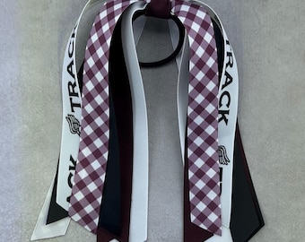 May include: A burgundy and white gingham hair bow with black and white ribbons. The ribbons have the text "TRACK" printed on them.