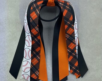 May include: A hair tie with a black elastic band, featuring ribbons in black, orange, and white. The design includes a baseball pattern and plaid accents. The ribbons are knotted at the top. The hair tie is suitable for various hairstyles.