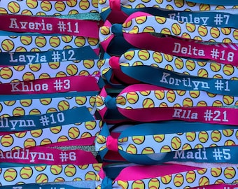 May include: A collection of hair bows featuring a softball pattern on white ribbon, accented with solid pink and teal ribbons. Each bow is personalised with a name and number in white text, including Averie #11, Finley #7, Layla #12, Delta #18, Khloe #3, Kortlyn #1, Ilynn #10, Ella #21, Addilynn #6, Madi #5, Kori #2, and Beretta #8.