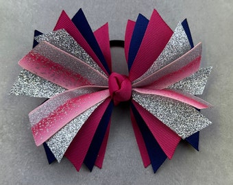 May include: A large, multi-layered bow made of pink, silver glitter, and navy blue ribbon. The bow is tied with a dark pink ribbon and has a silver glitter finish.