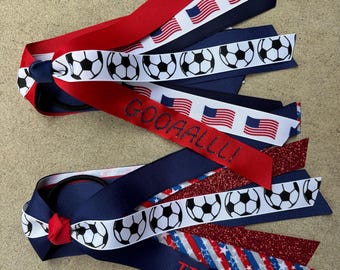 May include: Two hair ties featuring layered ribbons in red, white, and blue. One tie displays footballs, American flags, and the text GOOAALLL!. The other features footballs, red glitter, and a blue ribbon with the red embroidered text USA.