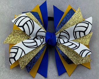 May include: A blue, gold and white cheer bow with a volleyball pattern. The bow is made with glitter ribbon and has a blue elastic band.