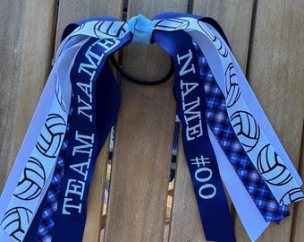Puede incluir: Una goma para el pelo azul y blanca con temática de voleibol. La goma para el pelo presenta cintas con las palabras "TEAM NAME" y "NAME #00" impresas. Las cintas son de varios tonos de azul y blanco, con gráficos de voleibol.