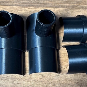 May include: Four black 3D-printed plastic components consisting of two T-shaped connectors with cylindrical ports and two matching semi-cylindrical curved sleeves. Each piece features a smooth finish and a rigid structure designed for assembly.