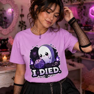 May include: A light purple t-shirt featuring a graphic of a sad ghost sitting next to a purple twenty-sided die. The design includes purple flames, a small skull, and the text I DIED in bold white letters with a black outline on the front.