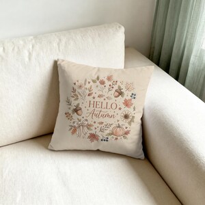 Op de afbeelding: Een vierkant beige sierkussen met een centrale kransillustratie van herfstelementen zoals eikels, esdoornbladeren, een pompoen, een strik en wilde bloemen. De tekst "HELLO Autumn" is in het midden in een bruin lettertype gedrukt.