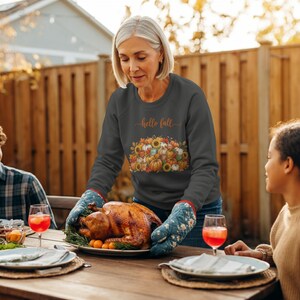 Op de afbeelding: Een donkergrijs sweatshirt met lange mouwen en de tekst Hello Fall boven een grafische illustratie van diverse pompoenen, zonnebloemen en herfstbladeren. Het sweatshirt wordt gedragen met blauwe ovenwanten met een bloemenpatroon.