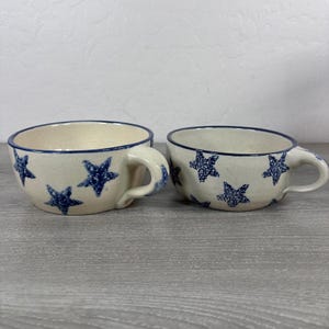 Może przedstawiać: Dwa kremowe kubki ceramiczne o nakrapianej fakturze z wzorem ciemnoniebieskich gwiazd. Każdy kubek ma zaokrąglony korpus, pojedyncze ucho z boku oraz ciemnoniebieski brzeg. Wzór jest spójny na obu elementach, prezentując ręcznie wykonany wygląd z gładkim wykończeniem i szerokim otworem.