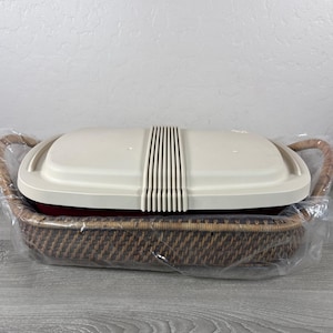 May include: A rectangular red casserole dish with a cream-colored plastic lid featuring a central ribbed design. The dish is nestled inside a woven brown wicker basket with integrated handles, all encased in a clear plastic protective wrap.
