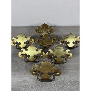 Può includere: Un set di sei maniglie per cassetti in ottone, ciascuna caratterizzata da una piastra posteriore ornamentale e simmetrica con intagli decorativi e una maniglia a arco oscillante fissata a due piccoli perni metallici.