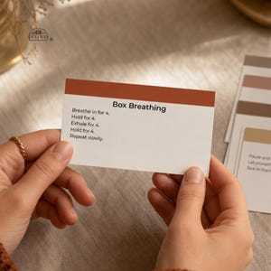 May include: A rectangular white card with a rust-colored top border featuring the black text Box Breathing followed by instructions: Breathe in for 4, Hold for 4, Exhale for 4, Hold for 4, Repeat slowly. The card is held against a neutral background.