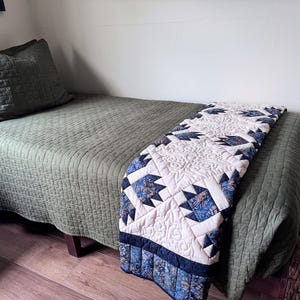 May include: A twin bed covered with a solid olive green quilted bedspread and a matching pillow sham. A decorative quilt runner is draped across the foot of the bed, featuring a white background with blue and brown floral bear paw patterns.