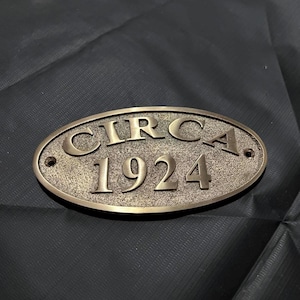 Könnte beinhalten: Ein ovales, bronzefarbenes Metallschild mit einer texturierten, körnigen Oberfläche. Das Schild zeigt erhabene, glatte Schriftzüge mit dem Wort "CIRCA" in der oberen Zeile und "1924" in der unteren Zeile. Es gibt zwei kleine Befestigungslöcher.