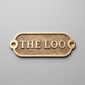 Könnte beinhalten: Ein rechteckiges Messingschild mit abgerundeten Enden und zwei Schraublöchern, das eine texturierte Oberfläche und den erhabenen, polierten Schriftzug THE LOO in einer klassischen Serifenschrift aufweist. Das Metall hat einen warmen Goldton.