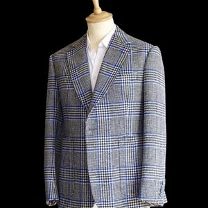 Peut inclure: Un blazer à motif écossais bleu, noir et blanc avec un revers cranté, deux poches avant et une fermeture à un bouton. Le vêtement est présenté sur un mannequin portant une chemise blanche à col rigide sous la veste.