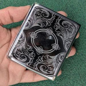 Può includere: Un portasigarette rettangolare in metallo con cornice in argento lucido e pannello frontale nero caratterizzato da intricati motivi floreali e volute bianche incise. L'astuccio presenta un lato incernierato e una finitura metallica dettagliata.