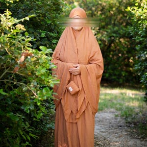 May include: A solid terracotta-colored prayer garment featuring a loose-fitting, floor-length robe with an integrated head covering. The ensemble includes a small, matching rectangular pouch with a wrist strap and a white label with black text.