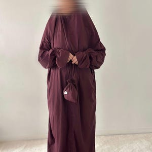 以下が含まれることがあります： 深いバーガンディ色の床まで届く長さのアバヤ。袖口はギャザーの入ったゴム仕様で、中央にはお揃いの巾着袋を持っています。この衣服は無地の滑らかな生地で作られており、床までまっすぐに垂れ下がっています。