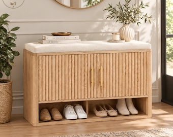 May include: A light wood shoe storage bench with a cream-colored upholstered seat, two fluted cabinet doors featuring vertical gold handles, and an open bottom shelf containing two pairs of loafers and two pairs of white sneakers.
