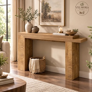 May include: A rectangular light brown wooden console table with thick slab legs. On the surface sits a white vase with green branches, a small dark bowl, a candle in a glass holder, a book, and a bowl of spheres. A woven basket sits below.
