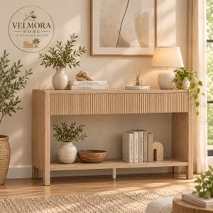 May include: A light wood console table with a fluted drawer front and an open bottom shelf. The table holds a ceramic vase with greenery, books titled Kinfolk and At Home, a table lamp with a textured base, and a wooden bowl.