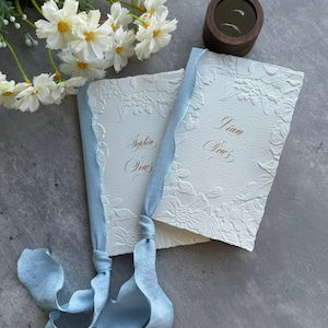 Può includere: Due libretti per i voti in carta bianca testurizzata con bordi floreali in rilievo e calligrafia dorata che riporta Sophia Vows e Liam Vows. I libretti sono legati insieme da un nastro di seta azzurro sfilacciato, accompagnati da fiori bianchi e un portafedi in legno.