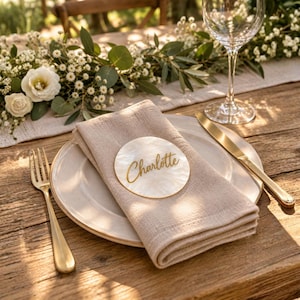 Könnte beinhalten: Ein rundes, weißes Platzkärtchen aus Perlmutt mit dem in goldener Schreibschrift geschriebenen Namen Charlotte, das auf einer gefalteten beigen Leinen-Serviette auf einem weißen Keramikteller liegt, begleitet von einer goldenen Gabel und einem Messer.