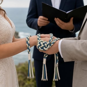 May include: A handfasting cord featuring braided strands in shades of blue, sage green, and cream. The cord is tied in a central decorative knot and features white woven collars at the ends, finished with long, loose tassels and silver-toned beads.