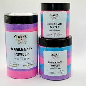 May include: Three cylindrical containers of Clarks & Chaos Bubble Bath Powder in Sweet Cheeks scent. The powder is layered in pink, blue, and purple. Labels indicate sizes of 14oz for the large jar and 8oz for the two smaller jars, with black lids.