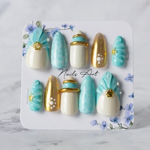 Peut inclure: Un ensemble de dix faux ongles sur une carte de présentation marquée Nails Art. Les motifs présentent un mélange de couleurs crème, turquoise et or métallique, avec des coquillages en relief, des étoiles de mer dorées et des bandes dorées.