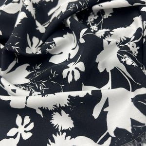 Peut inclure: Un morceau de tissu présentant un motif floral contrasté en noir et blanc. Le design se compose de grandes fleurs et de feuillages blancs stylisés imprimés sur un fond noir uni, avec un bord effiloché visible le long de la bordure inférieure.
