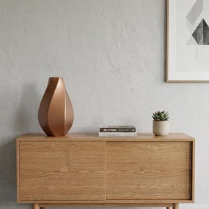 May include: A metallic copper-coloured vase with a faceted, geometric teardrop shape. The vase features a smooth, matte finish and a narrow opening at the top, standing as a singular decorative object on a light-toned wooden sideboard.