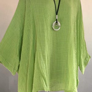 May include: A lime green loose-fitting tunic top with a textured fabric finish, featuring a wide round neckline and three-quarter length sleeves. A black cord necklace with a circular silver-toned pendant rests against the front of the garment.