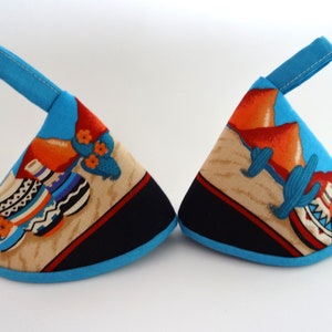 May include: Two blue pot holders with a southwestern design. One pot holder features a cactus and orange mountains, the other features a cactus and pottery. Both pot holders have a blue trim.