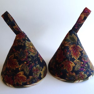 May include: Two dark blue fabric pot holders with a fall leaf and berry print. The pot holders have a loop handle and a cream and red trim.