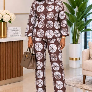 May include: A two-piece set consisting of a long-sleeved button-down shirt and matching straight-leg trousers. The garment features a dark brown fabric with a repeating white circular tie-dye pattern. A solid brown structured handbag is held.