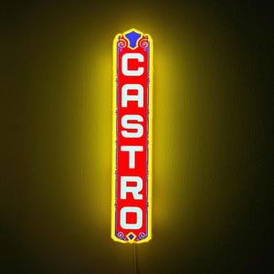 May include: A vertical rectangular wall sign with a bright yellow glowing border. The center features a red background with the word CASTRO written in bold white capital letters. The sign includes blue decorative scrollwork at the top and bottom.