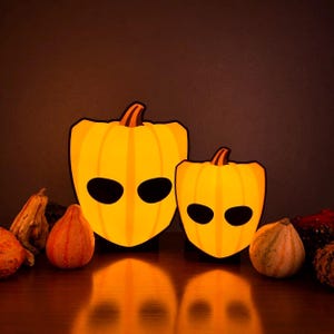 May include: Two illuminated pumpkin-shaped lamps with smooth yellow surfaces, dark brown stems, and two oval black eye cutouts. The lamps are positioned among several small, natural, multi-coloured gourds on a flat surface with a dark background.