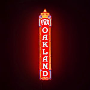 May include: A vertical rectangular neon sign featuring the word FOX at the top and OAKLAND written vertically below in white capital letters. The sign has a bright red background with yellow and blue decorative borders and a glowing light at the top.
