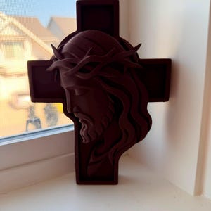 May include: A dark maroon, three-dimensional wall hanging featuring a sculpted profile of a face wearing a crown of thorns, layered over a simple cross shape. The object has a matte finish and displays a side view of the head and hair.