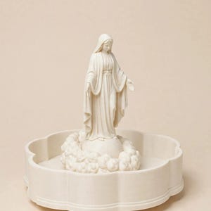 May include: A cream-coloured figurine of a standing figure wearing a long robe and head covering, positioned atop a spherical base surrounded by sculpted clouds, all resting inside a circular, scalloped-edge tray of the same matching colour.