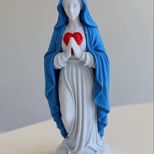 Puede incluir: Una figura blanca que viste un velo azul y una t&uacute;nica larga blanca, sobre una base circular. Las manos est&aacute;n juntas frente a un coraz&oacute;n rojo y lleva una corona amarilla con puntas en forma de estrella sobre la cabeza.