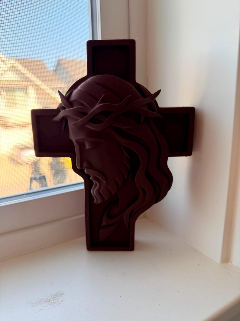 May include: A dark maroon, three-dimensional wall hanging featuring a sculpted profile of a face wearing a crown of thorns, layered over a simple cross shape. The object has a matte finish and displays a side view of the head and hair.