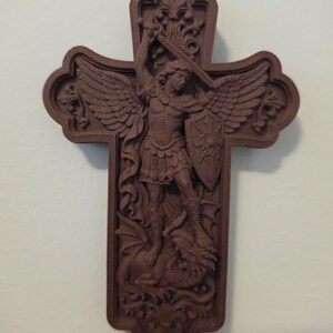 May include: A brown, cross-shaped wall plaque featuring a detailed relief carving of an angel with wings, holding a sword and shield, standing over a dragon. The design includes ornate scrollwork at the top and bottom of the cross frame.