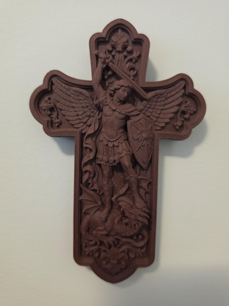 May include: A brown, cross-shaped wall plaque featuring a detailed relief carving of an angel with wings, holding a sword and shield, standing over a dragon. The design includes ornate scrollwork at the top and bottom of the cross frame.