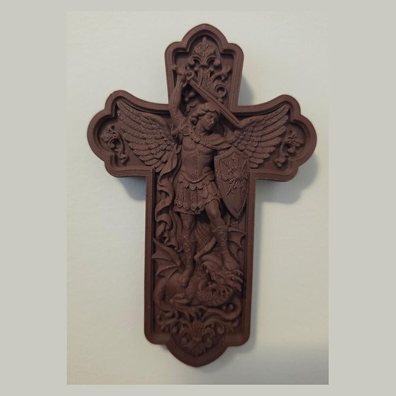 May include: A dark brown wall cross featuring a relief carving of a winged figure in armor holding a sword and shield while standing over a dragon. The cross has an ornate, scalloped edge design with intricate scrollwork details throughout.
