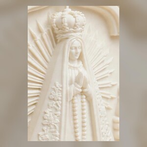 Puede incluir: Una escultura en relieve de color crema que representa una figura con corona y una t&uacute;nica larga y fluida con patrones florales. La figura tiene las manos juntas en oraci&oacute;n, con un rosario colgando al frente y un halo radiante detr&aacute;s.